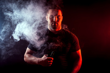 A young stylish man smoker in a black T-shirt is holding a    vape in his hand, and around and a big cloud of smoke from vape on a dark isolated background