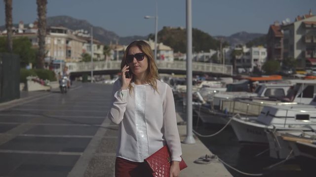 Positive business woman in sunglasses walking and talking on the phone on the background pier of the sea