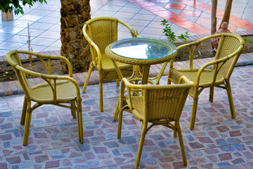 Wicker furniture: 4 chairs and a glass table, stand outdoors on a multicolored paving slab.