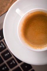 Cup of coffee on a laptop keyboard. Work place modern. Laptop (notebook) with cup of coffee and notepad with pen on old wooden table.