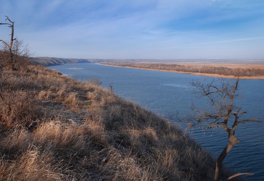 Autumn landscape yellow dry grass bare trees on the hill and the wide river with forest on background of blue sky with white clouds