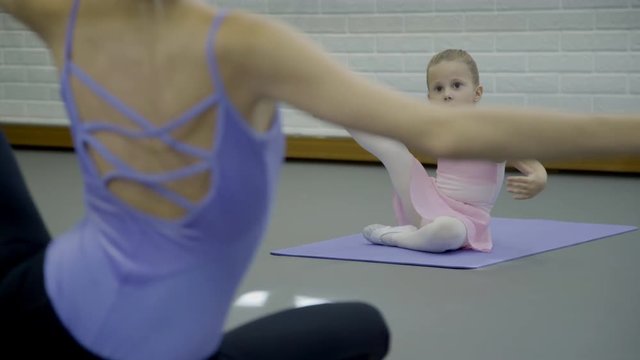 The prima ballerina teaches girl how to stretch the leg sideward in the modern bright studio. They sit on the perivinke mats on the floor and raised the feet. The little dancer in pink dress tries to