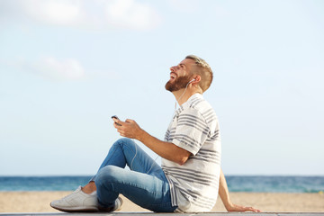 happy young man sitting by beach listening to music with mobile phone and earphones