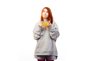 A young red-haired woman in a gray sweatshirt is sad because she fell ill with a grip and drinks from a large transparent cup an antiviral drug with a vitamin with a white isolated background