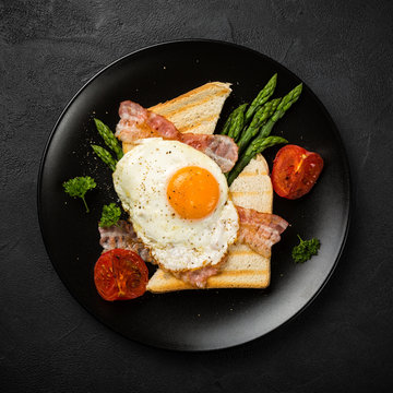 Breakfast or lunch with Fried egg, bread toast, green asparagus, tomatoes and bacon on black plate. Top view.
