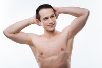 Fototapeta premium Sporty figure. Positive nice athletic man smiling and looking aside while holding hands behind his head