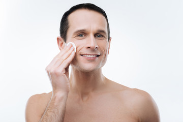 Male beauty. Cheerful joyful handsome man smiling and caring about his skin while cleaning it with a cotton pad