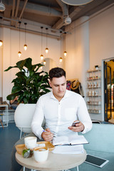 The stylish young businessman in a white shirt works sitting in cafe. Freelance. The man makes entries in a notebook.