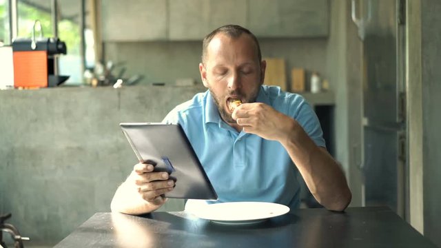 Young man with tablet eating breakfast sitting by table in kitchen at home
