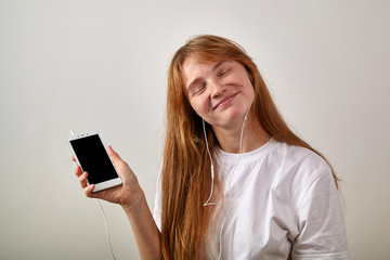 Fototapeta premium Headshot of young red-headed girl with freckles holding phone with earphones plugged in as if listening music, eyes closed, smiling