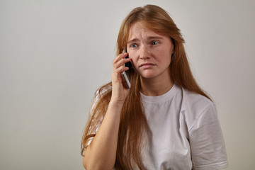 Studio indoor portrait of young red-headed girl with freckles holding her phone next to her ear and talking with sad expression on her face