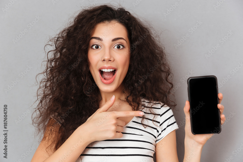 Sticker Close up photo of optimistic woman with ring in nose pointing index finger like advertising her smartphone meaning good choice against grey wall