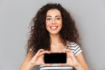 Fototapeta premium Optimistic woman with ring in nose holding her mobile phone meaning good choice smiling on camera while isolated against grey wall close up