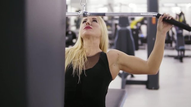 Attractive young blond caucasian woman with sporty body training hard on a lat pull-down mashine at the gym. Healthy lifestyle concept. Rotation footage