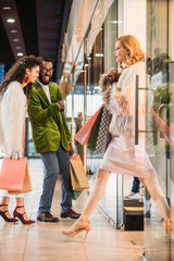 side view of stylish woman holding shopping bags and walking while smiling african american couple standing behind in mall