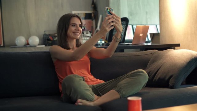 Happy woman taking selfie photo with cellphone sitting on sofa at home

