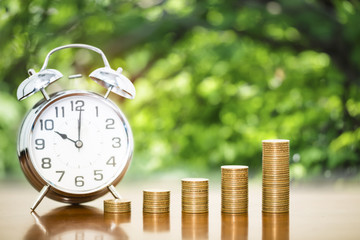 Money coin stack arranged as a graph on wooden table and alarm clock with blur nature background, concept of time to money growth