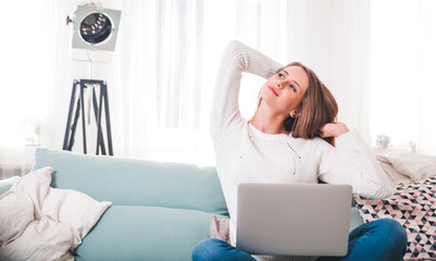 Woman at home sitting on sofa and stretching while working with laptop computer