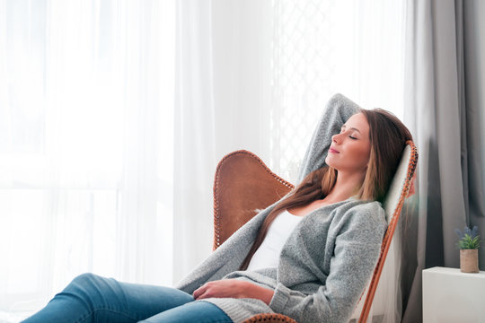 Woman at home sitting on modern chair near window relaxing in living room