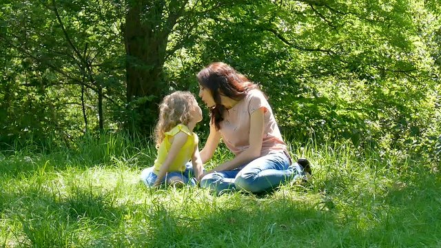 Happy Family Sitting on the Green Grass in the Park. Little Child Daughter Hugging and Kissing her Mother Outdoors During Sunny Day.