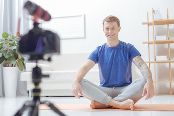 Cheerful person. Calm friendly handsome blogger sitting on the yoga mat in a lotus pose and smiling while waiting for his online lesson to start