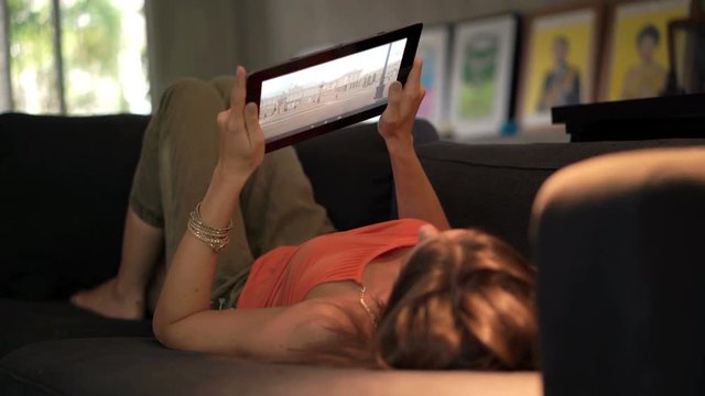 Woman browsing photos on tablet lying on sofa at home
