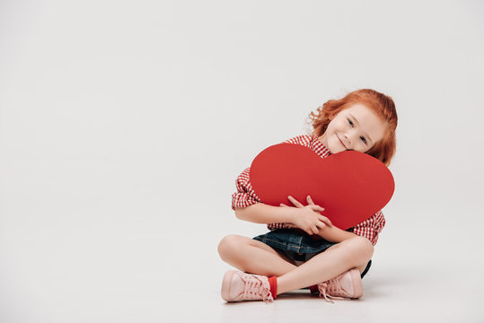 adorable little child hugging red heart symbol and smiling at camera isolated on grey