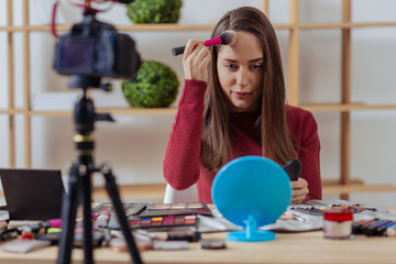 Attentive blogger. Clever enthusiastic young fashion blogger looking attentive while sitting in front of a mirror and putting on amazing facial powder