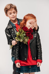boy presenting roses bouquet to his little girlfriend and covering her eyes with hand isolated on grey