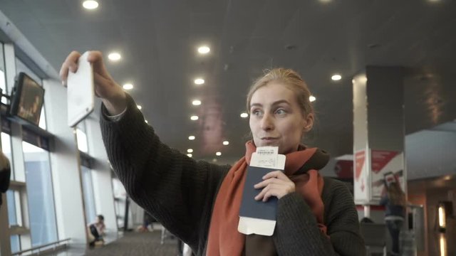 Happy young woman, holding French passport and boarding pass, doing selfie in the international airport near flight information board. Travel or vacation concept