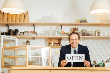 Dreams comes true. Busy satisfied perspective owner standing in his shop by the table showing the plate and smiling.