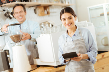 Calculate costs. Pleasant cute young woman in a pinafore standing near the kitchen smiling and using the calculator.