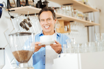 Fototapeta na wymiar Take and relax. Satisfied joyful gay man standing in the kitchen smiling and giving a cup of coffee.