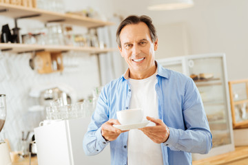Natural coffee. Handsome stylish smiling man standing in the kitchen looking straight and holding a cup of coffee.
