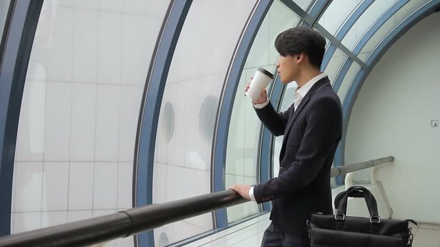 Male Asian man in jacket drinks coffee and looks at window on rain. Korean man near semicircular glass holds closed glass with drink and waits.