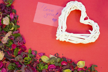 The concept of St.Valentine's Day with a woven heart and a love note, red background with a corner of rose petals and leaves top view, copy space