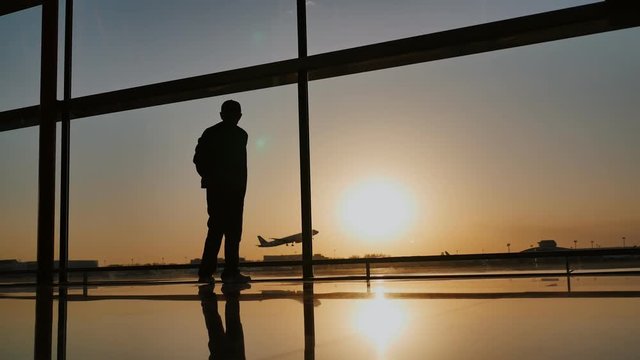 Silhouette of a tourist guy watching the take-off of the plane standing at the airport window at sunset in the evening. Travel concept, people in the airport.