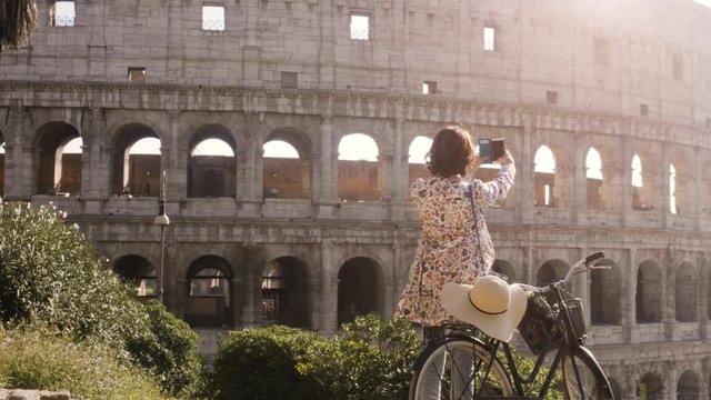 Beautiful young woman in colorful fashion dress alone on hill with bike taking pictures of colosseum in Rome with smartphone at sunset attractive girl with elegant straw hat