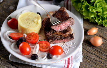 Beef steak with mashed potatoes and salad of cherry tomatoes, dried olives and white onions.