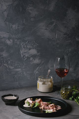 Vitello tonnato italian dish. Thin sliced veal with tuna sauce, capers and coriander served on black plate, glass of rose wine and ingredients above over gray texture kitchen table.