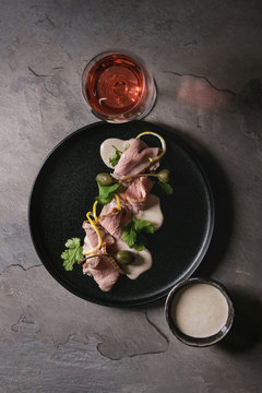 Vitello tonnato italian dish. Thin sliced veal with tuna sauce, capers and coriander served on black plate with glass of rose wine over gray texture kitchen table. Top view, space