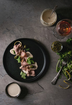 Vitello tonnato italian dish. Thin sliced veal with tuna sauce, capers and coriander served on black plate, glass of rose wine and ingredients above over gray texture kitchen table. Top view, space