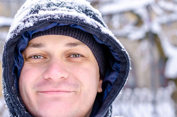 Young funny happy stylish smiling man dressed winter clothes frozen ski jacket looks at the camera at background of falling snow of the winter city street outside hurdel winter snowfall