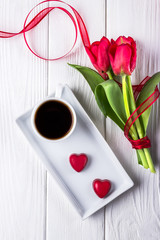 Black coffee in a white cup, red tulips and two red hearts candy on a white background. Top view, flat lay. Happy Valentine's Day!