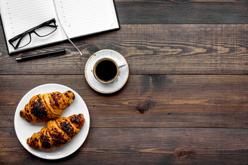 Work at breakfast. Coffee and croissants near notebook and glasses. Dark wooden background top view copy space