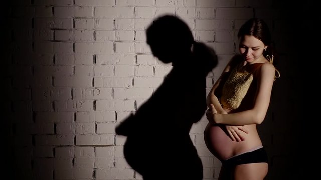 young pregnant woman is standing in background of white brick wall in a light of a soffits
