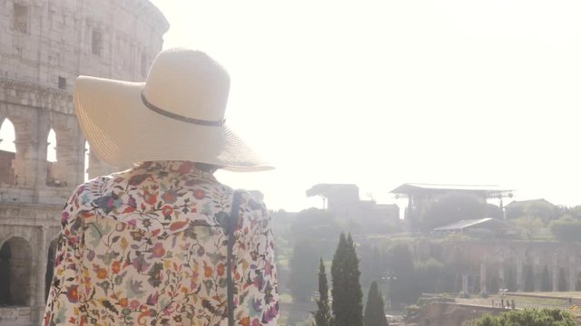 Beautiful young woman in colorful fashion dress alone in front of colosseum in Rome at sunset attractive girl with taking off elegant straw hat