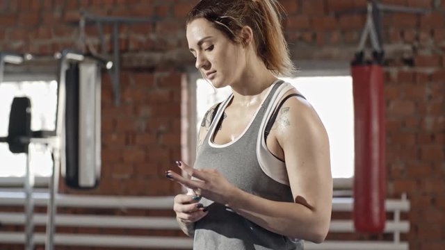 Young sweaty woman with tattooed body wrapping her fingers with tape and cutting it with teeth while training hard in gym
