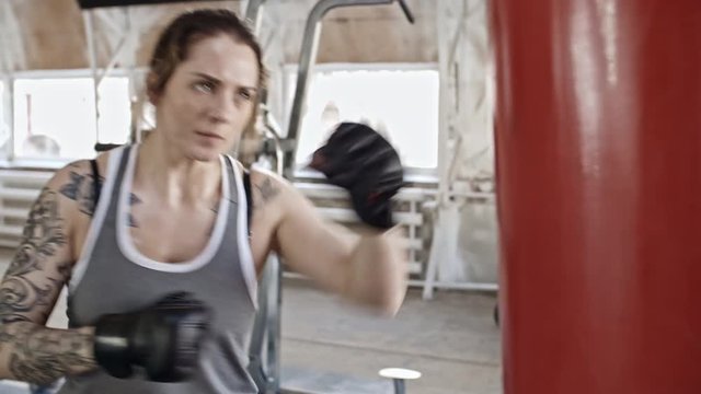 Handheld camera shot of young female boxer in gloves hitting punching bag while training in gym