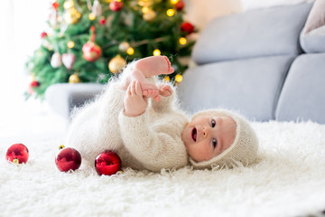 Little baby boy in white knitted onesie, playing with and opening presents at home